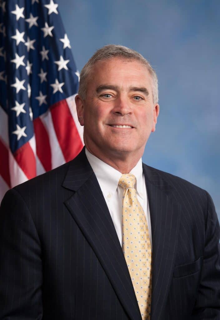 Prominent man in suit with American flag in background.
